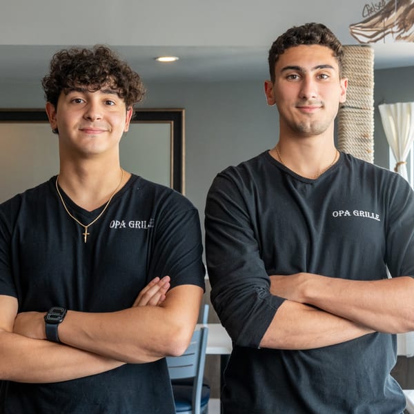 Two young men in black Opa Grille shirts stand with arms crossed, smiling at the camera.