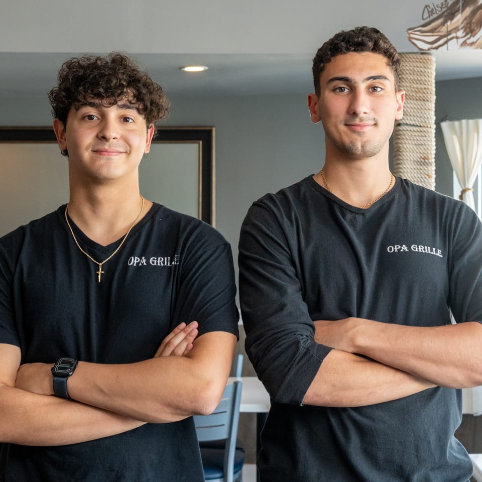 Two young men in black Opa Grille shirts stand with arms crossed, smiling at the camera.