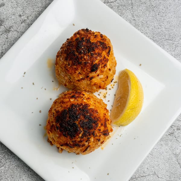 Two golden-brown Greek crab cakes with a lemon wedge and seasoning on a white plate.